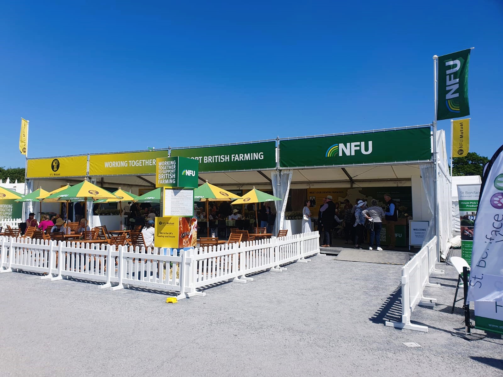 NFU Farming trade stand at Devon County. Outside the marquee are white fences with an area for guests to relax and sit under the table umbrellas. 