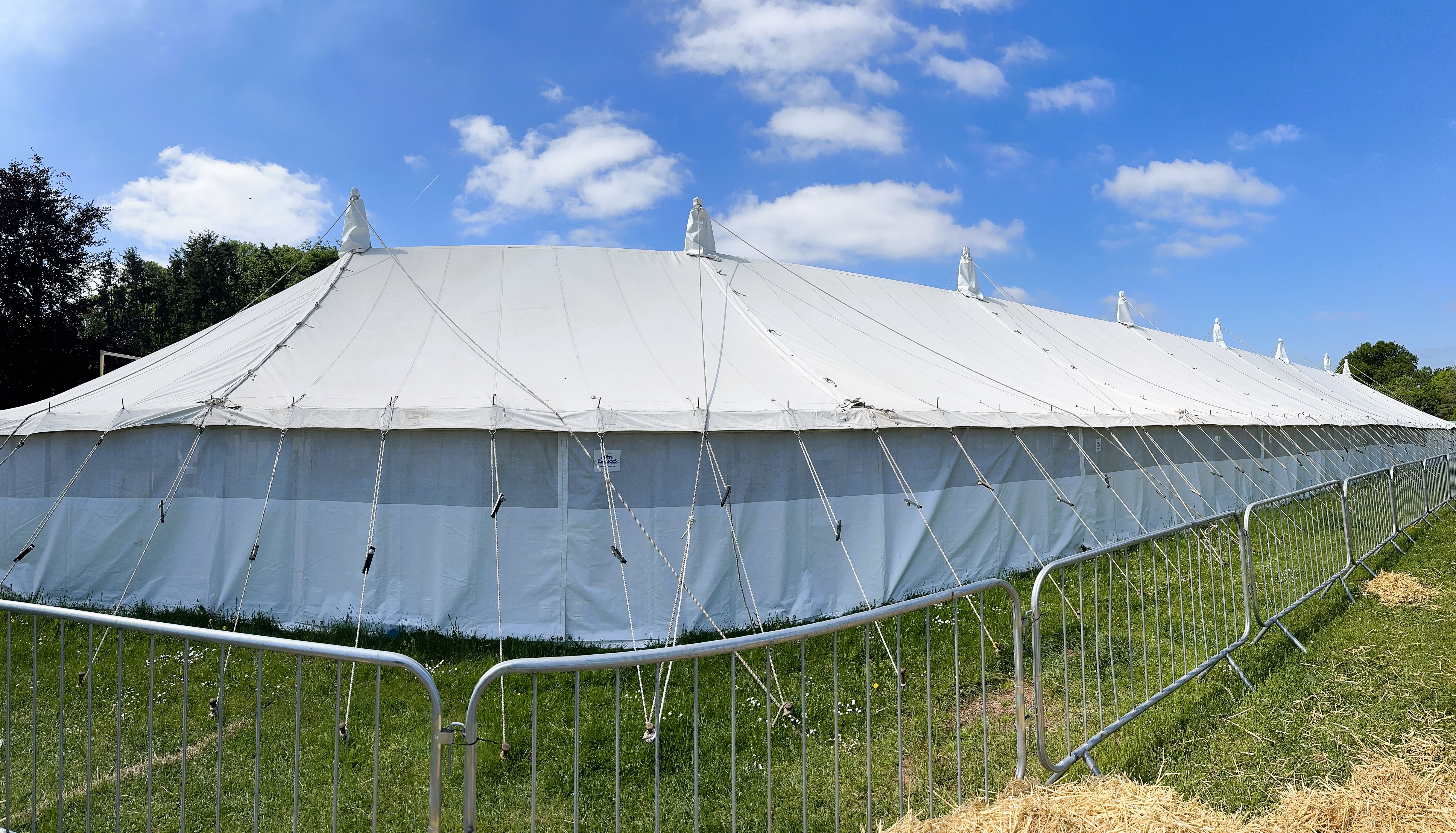 A traditional marquee structure, pegged into the grass and surrounded by metal pedestrian barrier. A blue sky filled with clouds. 