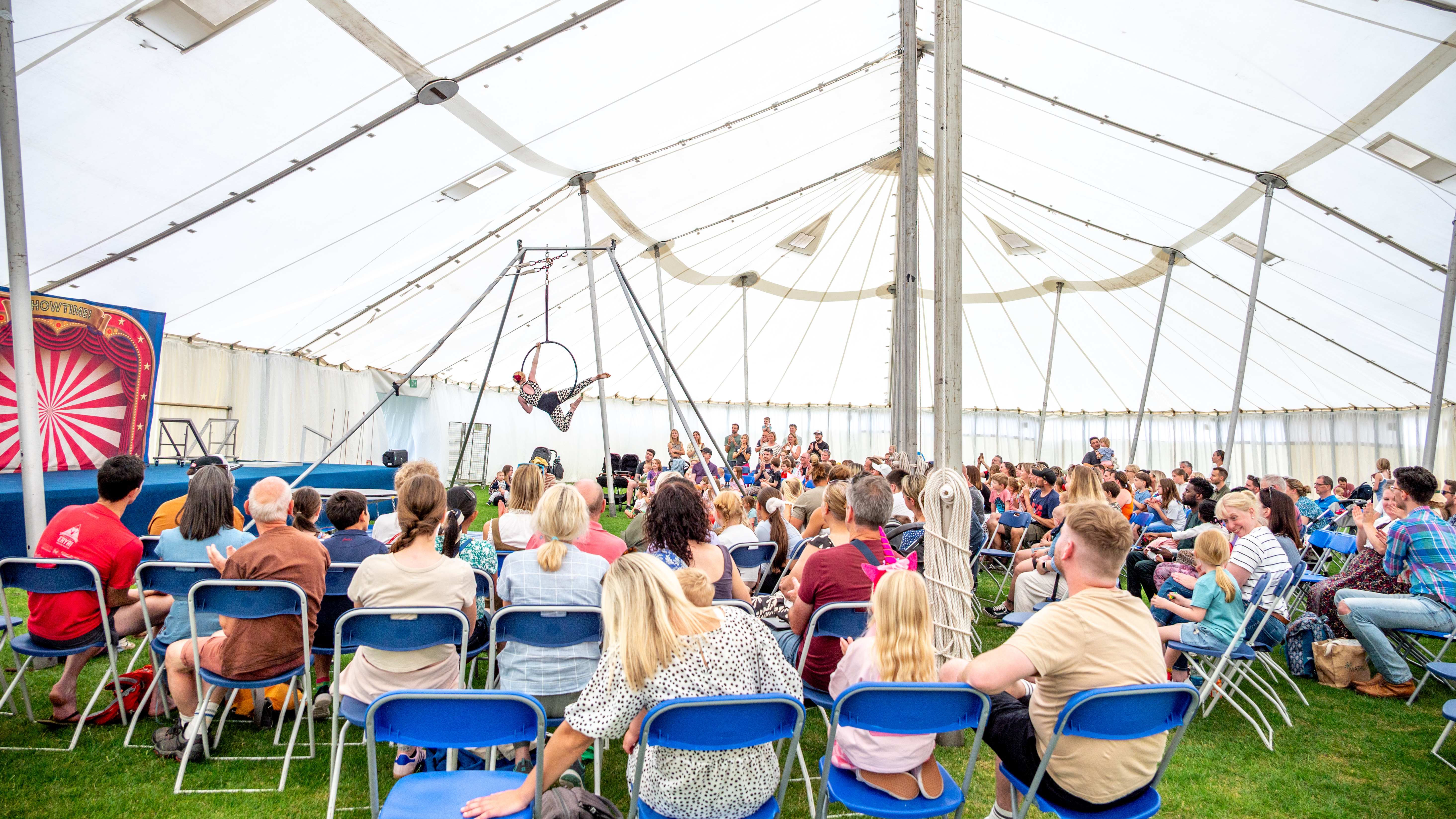 A circus performer is dangling in a hoop which is hanging from the top of a traditional marquee. Spectators are enjoying the circus show. 