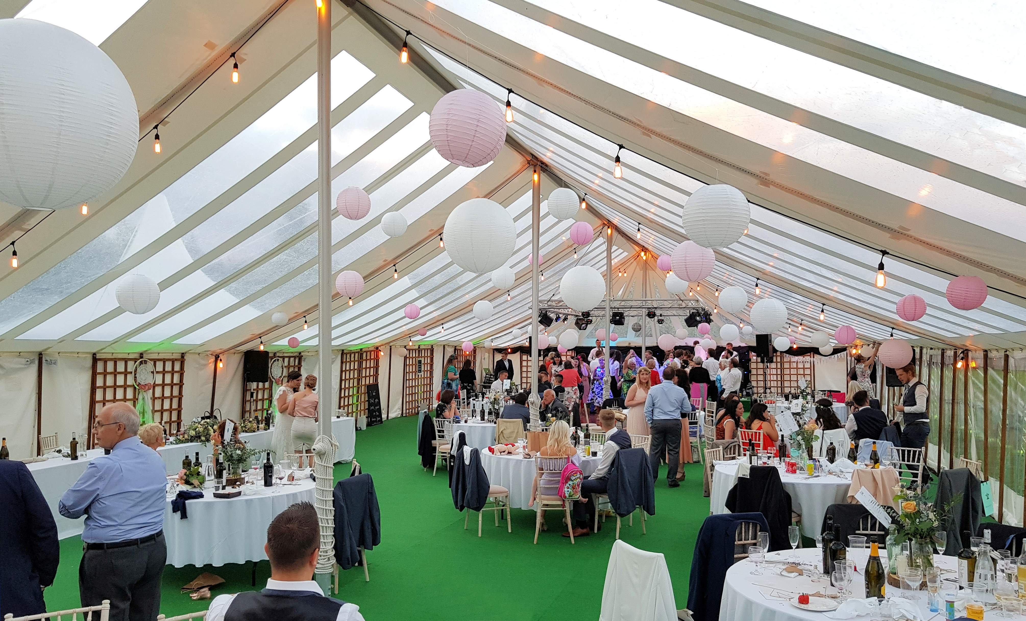 A wedding gathering inside a Traditional Marquee with semi clear roofs and walls with decorative hanging paper lanterns