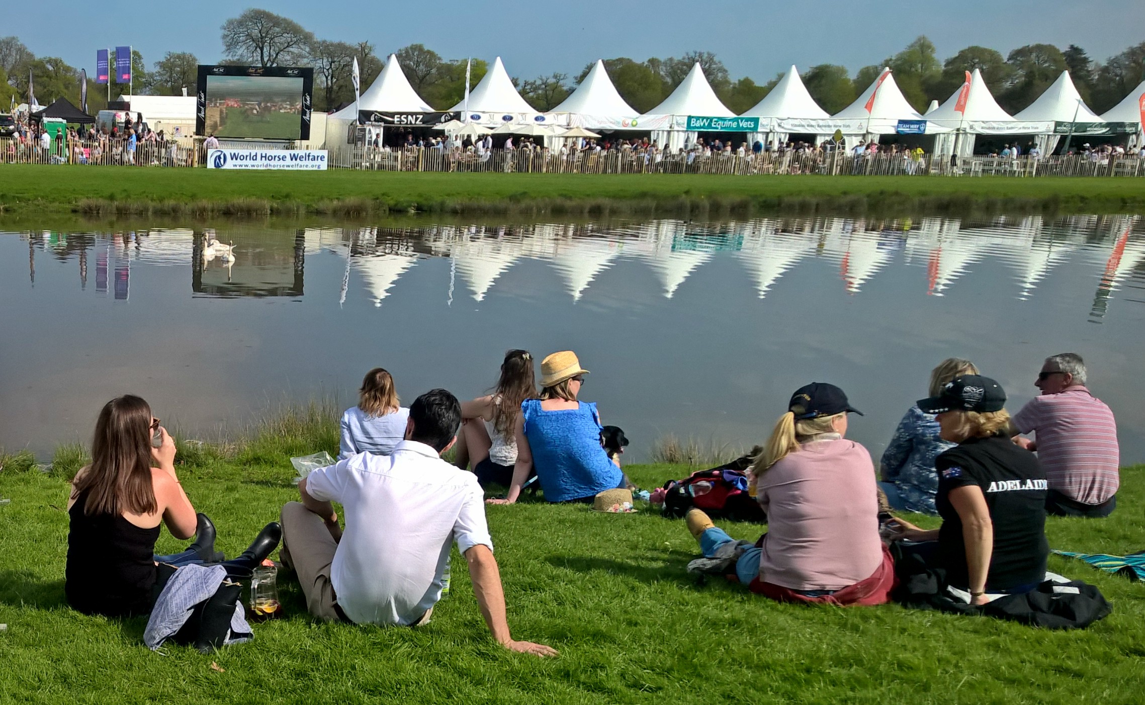 Danco marquee hats reflect on the water at Badminton Horse Trials. People are sat on the grass next to the lake enjoying the event.
