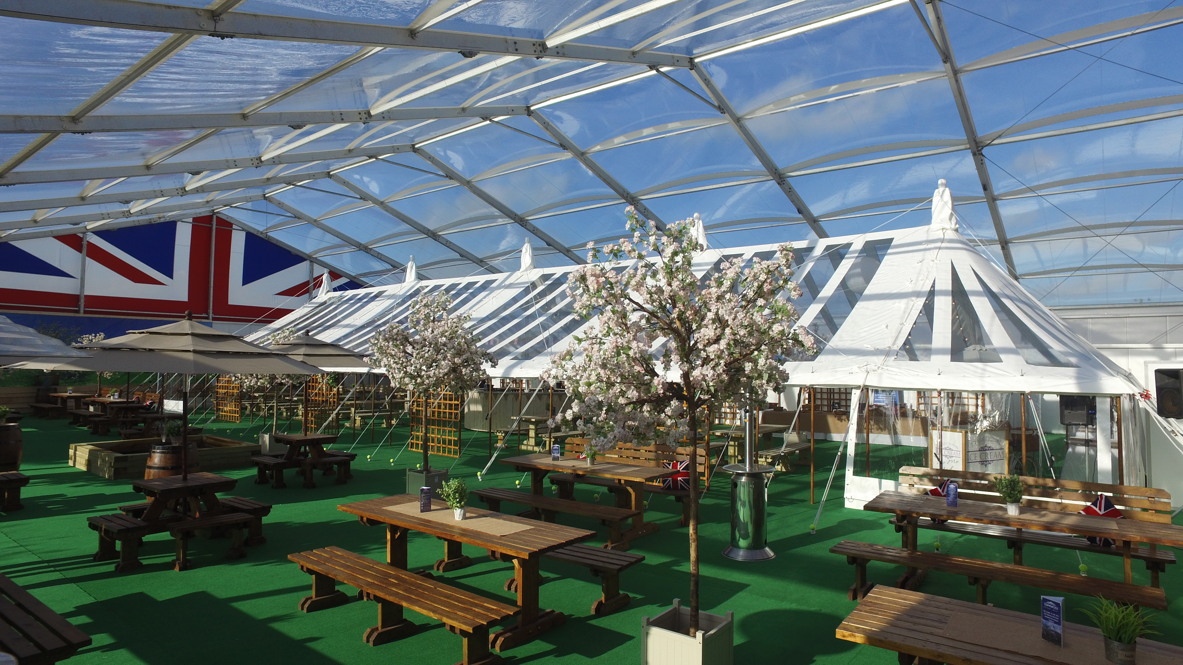A hospitilaty marquee. A traditional tent is inside a clear span marquee which is also filled with wooden picnic style table and chairs, decorateive trees and plants,. 